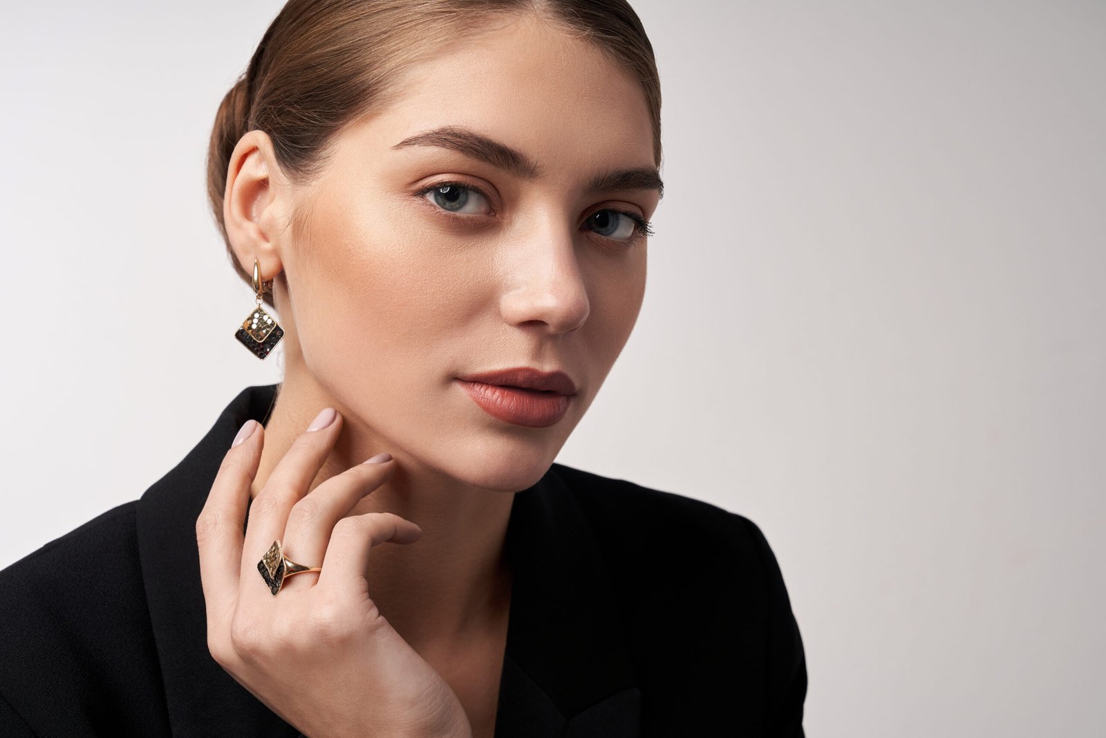 Close up portrait of young beautiful female model in formal suit presenting golden and silver earrings and ring. Woman with perfect makeup posing in studio, looking at camera, isolated on white.